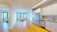 This thoughtfully designed single-floor unit showcases a clean, contemporary aesthetic with an open-concept layout that seamlessly connects the living area and kitchen. Warm-toned hardwood flooring runs throughout the main living spaces, lending a sense of continuity and warmth to the interior.

The living area is bright and airy, anchored by a large, multi-panel window that frames views of the surrounding urban landscape and invites ample natural light. A wall-mounted HVAC unit sits beneath the window, providing efficient climate control.

The kitchen is positioned along one side of the open floor plan and is outfitted with a full suite of stainless steel appliances, including a French door refrigerator, gas range, over-the-range microwave, and dishwasher. Flat-front white cabinetry extends from floor to ceiling, maximizing storage, while a subway tile backsplash and clean countertops complete the modern look. Recessed linear lighting above the kitchen zone adds a polished architectural detail.

The bathroom features neutral mosaic tile flooring, a wall-mounted vanity with a vessel sink, a large mirror with built-in lighting, and a window that introduces natural light. The overall design of the space is clean, minimal, and functional, making it well-suited for modern urban living.