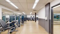 Located at 400 S 40th St in Philadelphia, Pennsylvania, this fitness facility offers a comprehensive, single-floor layout designed to support a wide range of workout activities. The space is thoughtfully organized into distinct functional zones, all finished with light hardwood-style flooring and accented by a two-tone gray and white wall design that gives the interior a clean, modern aesthetic.

The main exercise room is generously sized and outfitted with a lineup of Matrix-brand strength training machines and a stationary cycling bike. Full-length wall mirrors amplify the sense of space and allow users to monitor their form during workouts. A dedicated free weights area features a well-stocked rack of dumbbells and kettlebells, providing options for a variety of strength and conditioning exercises.

A separate studio area is appointed with wall-mounted ballet barres and large framed mirrors, making it well-suited for barre classes, stretching, or movement-based training. Adjacent to this, a functional training zone houses stability balls in multiple sizes displayed on wall-mounted racks, medicine balls, a folding mat, and additional accessories for dynamic exercise routines.

Throughout the facility, a drop ceiling with integrated fluorescent lighting ensures consistent, bright illumination. The entry area features glass-panel doors, a planted accent, and clear signage, contributing to a welcoming and professional environment.