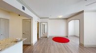 This single-floor residence showcases a cohesive design defined by light wood-style flooring, crisp white walls, and crown molding that runs throughout the main living areas. The open living space is generously proportioned, with a ceiling fan, flush-mount lighting, and a distinctive arched doorway that adds architectural character to the layout. Natural light filters in through a window fitted with blinds, enhancing the bright and airy atmosphere.

The kitchen is well-appointed, featuring granite countertops, gray cabinetry, a stainless steel undermount sink with a modern faucet, and a full suite of stainless steel appliances including a range, microwave, and dishwasher. A dedicated pantry or utility closet provides additional storage directly off the kitchen.

A hallway connects the living area to the private spaces, including a bathroom outfitted with a wide vanity, light-colored countertop, gray cabinetry with brushed nickel hardware, and a large mirror illuminated by a row of globe lights. The walk-in closet offers substantial storage capacity, with wire shelving lining three walls and carpeted flooring underfoot.

Overall, the space presents a clean, functional aesthetic with consistent finishes throughout.