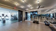 This well-appointed fitness center is designed to accommodate a wide range of workout routines within a clean, contemporary setting. The space is divided into distinct functional zones, seamlessly connected through glass-panel partitions and sleek folding doors. A dedicated cardio room, visible through the glass wall, houses treadmills and additional exercise equipment, providing a focused environment for machine-based training.

The main exercise studio features smooth hardwood-style flooring, ideal for group fitness classes, stretching, or freestanding workouts. A large flat-screen television is prominently mounted on the wall, offering entertainment or instructional content during sessions. Floor-to-ceiling mirrors line the walls, visually expanding the space while allowing users to monitor their form. Step platforms and other portable fitness accessories are neatly arranged on the floor, adding functional versatility.

Overhead, a combination of track lighting and ceiling fans ensures the space remains well-lit and comfortable during use. The overall ambiance is modern and energizing, with a neutral color palette, refined finishes, and thoughtful design details that create an inviting and professional fitness environment.