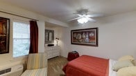 This single-floor space centers around a well-appointed bedroom designed for comfort and functionality. The room is finished with neutral walls that serve as a clean backdrop for its warm, decorative touches. A ceiling fan with an integrated light fixture provides both air circulation and ambient lighting overhead.

A large, ornately framed artwork depicting florals in rich reds and creams serves as a prominent focal point on the main wall, adding a sense of elegance to the space. The bedroom accommodates a full-sized bed dressed in a bold red comforter, complemented by a striped upholstered chair that introduces a relaxed, casual element.

An alcove off the main room functions as a vanity or dressing area, complete with a white multi-drawer dresser, a decorative lamp, and an ornate framed mirror that reflects light back into the space. The carpeted flooring runs throughout, contributing to the room's cozy, lived-in feel. Deep red curtains frame a window, tying the color palette together and adding warmth to the overall ambiance.