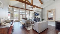 This single-story space showcases an impressive open-plan layout anchored by dramatic vaulted ceilings adorned with prominent exposed wood beams, lending a warm, rustic-modern character to the interior. Dark hardwood flooring runs throughout, providing a rich, cohesive foundation that complements the neutral gray and white color palette.

The main lounge area is generously sized, furnished with nailhead-trim sofas and accent chairs arranged around a wooden coffee table atop a patterned area rug. A fireplace with a classic white surround serves as a focal point on one wall, while a wall-mounted TV and decorative mirrors add visual interest. Ceiling fans with dark wood blades enhance the architectural detail above. Large multi-pane windows flood the space with natural light and offer views of the surrounding greenery.

An adjacent workstation area provides computer terminals for added functionality. The kitchen features gray shaker-style cabinetry, white subway tile backsplash, light quartz countertops, and a full suite of stainless steel appliances. A center island with a natural wood base and pendant lighting above adds workspace and style. A window above the sink allows natural light to brighten the kitchen throughout the day.