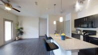 This single-story space showcases a thoughtfully designed open-concept layout that seamlessly connects the kitchen and living areas. The kitchen serves as a central focal point, anchored by a generous granite-topped island with a built-in sink and seating for bar stools, making it ideal for casual dining or entertaining. Sleek dark cabinetry provides a striking contrast against the light granite countertops, while two elegant pendant lights hang above the island, adding both functional illumination and a contemporary aesthetic touch.

The flooring throughout the main living area consists of dark hardwood-style planks, lending warmth and visual continuity to the space. White walls and a light-colored ceiling enhance the sense of openness and allow natural and artificial light to reflect evenly throughout. A ceiling fan is partially visible, contributing to the room's comfort and airflow.

Beyond the kitchen, the open living area offers a clean, versatile space with clear sightlines toward what appears to be a hallway leading to additional rooms. Louvered closet doors are visible in the background, hinting at practical storage solutions integrated into the layout. Overall, the space presents a clean, modern aesthetic with a well-balanced combination of functionality and style.