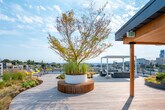 This rooftop terrace offers a sophisticated outdoor retreat elevated above the surrounding cityscape, accessible via elevator or stairwell. The deck is finished in warm-toned wood planking that extends generously across the space, creating an inviting platform for outdoor living and gathering. A standout architectural feature is the sleek white pergola with an adjustable louvered roof, positioned to frame sweeping panoramic views of the city skyline while providing flexible shade and shelter. Beneath it, a comfortable seating arrangement with upholstered outdoor sofas and armchairs creates an ideal setting for relaxation and socializing. Thoughtfully integrated landscaping adds a natural dimension to the space, with large circular and curved planter beds filled with ornamental grasses, seasonal plantings, and mature trees that soften the urban environment. The perimeter is enclosed by a glass-panel railing system that preserves unobstructed sightlines to the skyline and surrounding neighborhoods. Practical amenities include dedicated trash and recycling stations, a mounted fire extinguisher, and clearly marked building safety signage. The combination of refined materials, intentional design, and breathtaking views makes this rooftop terrace an exceptional amenity space.