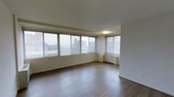 This single-floor space offers a clean and functional layout designed to maximize comfort and natural light. The main living area is a standout feature, characterized by a wide bank of large windows that span nearly the full length of one wall, flooding the room with abundant daylight and offering open views of the surrounding urban cityscape. The windows are fitted with horizontal blinds, providing flexible light and privacy control throughout the day.

The flooring throughout the space consists of smooth, medium-toned hardwood planks that contribute warmth and a polished aesthetic. The walls and ceiling are finished in a crisp, neutral white, creating a bright and airy atmosphere that feels open and inviting. A flush ceiling light fixture provides overhead illumination, complementing the natural light from the windows.

A radiator unit is positioned beneath the windows, ensuring efficient climate control during cooler months. The overall layout is open and unobstructed, offering significant flexibility for a variety of furniture arrangements and uses. The minimalist design aesthetic, combined with the generous natural light and urban outlook, makes this a practical and appealing space suitable for residential or professional occupancy.