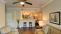 This single-story space showcases an open-concept floor plan that seamlessly integrates the living, dining, and kitchen areas into one cohesive environment. The living area features rich, dark hardwood flooring that extends throughout, providing a warm and grounded aesthetic. Crown molding trims the ceiling, adding a refined architectural detail, while a ceiling fan with a built-in light fixture serves as a functional centerpiece overhead.

The kitchen is positioned at the heart of the layout, accessible via a raised breakfast bar that accommodates two bar stools, making it an ideal spot for casual dining or conversation. Pendant lighting hangs above the bar in a trio of fixtures, casting a warm glow over the space. The kitchen is equipped with stainless steel appliances, including a microwave, and features light-toned cabinetry that contrasts with the darker flooring.

Adjacent to the kitchen, a dedicated dining area is visible, furnished with a dining table and chairs, and accented with decorative wall art and branch-style decor. The living area is furnished with neutral-toned seating and complemented by ambient table lighting, creating an inviting and well-balanced atmosphere throughout the space.