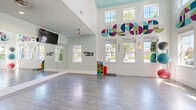 Located at 5400 Walkertown Landing Cir in Walkertown, NC, this energetic fitness studio is thoughtfully designed to inspire movement and wellness. The single-floor space is defined by dramatically high ceilings that lend an open, airy atmosphere, complemented by a bold, colorful geometric mural that wraps across the upper walls, creating a visually dynamic and motivating environment. Large windows on multiple walls flood the studio with abundant natural light and provide views of the surrounding greenery, enhancing the bright and uplifting ambiance. The open workout floor features smooth, light-toned hardwood-style flooring that provides an ideal surface for a wide range of fitness activities. A full wall of mirrors along one side expands the sense of space and supports form-checking during workouts. Dual wall-mounted flat-screen TVs and ceiling fans with integrated lighting ensure both comfort and entertainment throughout sessions. Equipment storage, including racks for stability balls and medicine balls, is neatly organized within the studio. A glazed partition wall separates an adjoining fitness area, maintaining visual connectivity while defining distinct workout zones. Motivational wall decals, colorful accent artwork, and seating benches near the entry complete the welcoming, purpose-driven design of this well-appointed studio space.