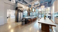 This expansive single-floor amenity space showcases a striking industrial-modern design, characterized by soaring ceilings with exposed ductwork and structural elements painted in a cool blue-grey palette. Polished concrete floors run throughout, lending a sleek, cohesive feel to the entire area. Floor-to-ceiling windows along the perimeter flood the space with abundant natural light and offer open views of the surrounding streetscape.

The open-concept layout is divided into several well-defined functional zones. The fully equipped kitchen features a large central island with bar seating, dark cabinetry, stainless steel appliances including a double wall oven, a full-size refrigerator, and a substantial stainless steel range hood — all illuminated by a collection of elegant pendant lights. Adjacent to the kitchen, a generous dining area accommodates a long farmhouse-style table surrounded by modern white chairs, set beside a sleek, linear built-in fireplace.

The lounge zone offers a large sectional sofa oriented toward a wall-mounted flat-screen television. A billiards area with a full-size pool table is defined by a glass panel railing, adding a contemporary touch. Accent lighting, curated artwork, and tasteful décor details complete the sophisticated yet relaxed ambiance of this multifunctional communal space.