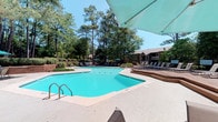 This outdoor amenity space centers around a generously sized freeform swimming pool with clear, aquamarine water and classic chrome entry rails. The surrounding deck provides ample room for lounging, with rows of chaise lounge chairs arranged to capture full sun exposure throughout the day. Several teal market umbrellas are positioned throughout the deck area, offering shaded retreats for those seeking relief from the sun. A raised wooden platform area to one side adds a tiered dimension to the layout, accommodating additional seating with a slightly elevated perspective of the pool.

The space is framed by a dense canopy of tall, mature pine and deciduous trees that provide a natural privacy screen and contribute to a tranquil, park-like atmosphere. Manicured shrubs and low landscaping line the perimeter, adding greenery and definition to the space. In the background, a residential building with a covered porch is partially visible, indicating this pool area serves as a shared community amenity. The overall ambiance is relaxed and inviting, combining functional outdoor recreation with a serene natural setting ideal for warm-weather enjoyment.
