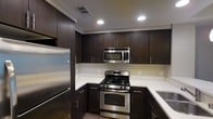This single-floor space showcases a thoughtfully designed interior with a strong emphasis on modern aesthetics and functional living. The kitchen is a standout feature, boasting rich dark espresso-toned cabinetry that extends floor to ceiling, providing ample storage throughout. Sleek white countertops offer a striking contrast against the dark cabinetry, creating a bold yet refined visual balance. The kitchen is fully equipped with a suite of stainless steel appliances, including a gas range with oven, an over-the-range microwave, and a full-size refrigerator, making it both stylish and highly functional. Recessed lighting is strategically placed throughout the kitchen ceiling, ensuring bright, even illumination across the workspace. The cabinetry hardware features a clean, minimalist bar-pull design that complements the overall contemporary style. The layout is designed to maximize efficiency, with an L-shaped counter configuration that allows for a smooth workflow. The space features consistent flooring that ties the interior together cohesively. Overall, the space exudes a modern, polished ambiance with attention to detail in both design and functionality.