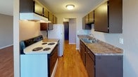 This single-floor unit offers a practical and well-organized layout across several distinct living areas. The living room is a standout space, featuring rich hardwood-style flooring that reflects natural light pouring in through large vertical-blind-covered sliding glass doors, offering views of the outdoors. A wall-mounted air conditioning unit and baseboard heating ensure year-round comfort. The open sightlines and textured walls give the living area a clean, functional character.

The galley-style kitchen is efficiently designed with dark wood cabinetry lining both sides, providing ample storage space. Laminate countertops, a double-basin sink, a coil electric range, and a full-size refrigerator complete the kitchen's practical setup. The hardwood-style flooring continues seamlessly from the living area into the kitchen, unifying the space.

The bedroom features soft, neutral carpet underfoot, a ceiling fan with light fixture, and a generously sized window dressed with vertical blinds, allowing for natural light and outdoor views. The room offers a calm and comfortable retreat.

The bathroom is cleanly appointed with mosaic tile flooring, a single-sink vanity with dark wood cabinetry, a wall-mounted mirror with overhead lighting, and a full bathtub and shower combination with white tile surround. Throughout the unit, neutral tones and textured ceilings create a cohesive, understated aesthetic.