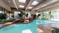 This impressive indoor aquatic facility offers a resort-like experience within a thoughtfully designed single-level atrium space. The centerpiece is a generously sized freeform pool with vivid turquoise water, its organic curves lending a natural, lagoon-like character to the environment. A striking rock formation water feature anchors one end of the pool, adding a naturalistic focal point that enhances the overall ambiance.

The architecture draws the eye upward to a series of large skylights set into a structured ceiling with exposed beams, flooding the interior with abundant natural light throughout the day. Floor-to-ceiling windows along the perimeter walls further reinforce the bright, open atmosphere while offering views to the surrounding exterior greenery.

Lush tropical plants and mature trees are strategically positioned throughout the space, creating a vibrant, garden-like atmosphere that softens the architectural lines and contributes to a tranquil, retreat-like feel. Tiled stone decking surrounds the pool, complementing the earthy tones of the rock feature. Seating areas with lounge chairs are visible along the edges, providing comfortable spots to relax. The combination of natural materials, generous daylighting, and verdant plantings makes this indoor pool facility both functional and visually inviting.
