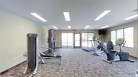 This fitness center offers a clean, functional environment designed to support a well-rounded workout routine. The space is laid out on a single level with a patterned carpet floor that adds a degree of comfort underfoot. Overhead, large rectangular panel lights provide bright, even illumination throughout the room, ensuring excellent visibility during workouts.

The equipment selection covers both strength and cardio training needs. A multi-station cable machine anchors the strength training area, accompanied by a weight bench for free weight exercises. On the cardio side, the space features a treadmill and an elliptical trainer, both positioned near the exterior wall. A wall-mounted flat-screen television is also present, allowing guests to stay entertained during their sessions.

A standout feature of this fitness center is the glass sliding door and adjacent windows along the far wall, which flood the room with natural light and offer views of the surrounding greenery outdoors. The neutral, light-toned walls contribute to an open and airy atmosphere. The overall ambiance is practical and welcoming, making it a convenient and comfortable space for daily exercise.