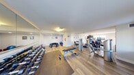 This fitness center offers a thoroughly outfitted exercise facility designed to support a wide range of workout routines. The space is laid out across a single floor with light wood-style flooring throughout, complemented by clean white walls and flush-mounted lighting that creates a bright, welcoming atmosphere. Protective foam matting is strategically placed in key workout zones for added comfort and safety during floor exercises.

The cardio section is well-stocked with multiple treadmills, elliptical trainers, upright and recumbent stationary bikes, and stair-climbing machines, each equipped with digital display consoles. Strength training is addressed through a selection of cable and resistance machines, adjustable weight benches, and a full rack of dumbbells lining one mirrored wall. The mirrors serve to visually expand the space and allow users to monitor their form during workouts.

A dedicated functional training area provides room for stretching, core work, and accessory exercises, with stability balls and additional free-weight equipment readily available. Natural light filters in through several windows, offering views of the surrounding outdoor environment and contributing to an energizing workout atmosphere. The space also includes a fire safety system, and access points connect to adjoining areas of the broader facility.