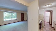 This two-story residence offers a straightforward and functional layout spread across two floors. The main level features an open living area finished with neutral-toned carpet and cream-colored walls, allowing natural light to filter in through a large window overlooking mature trees outdoors. A textured ceiling adds subtle character to the space. Adjacent to the living area, the kitchen is semi-partitioned and equipped with white cabinetry trimmed in warm wood tones, a double stainless steel sink set into a light-colored countertop, a range, and a full-size refrigerator. Ample upper cabinet storage and a dedicated shelf above the sink add to the kitchen's practicality. A staircase leads to the second floor, where multiple carpeted bedrooms are located, each featuring windows with views of surrounding greenery, closet access, and consistent neutral finishes. The bathroom includes a full tub and shower combination, a toilet, a single-basin vanity with a large mirror, and tile flooring. Throughout both floors, dark wood baseboards provide a consistent design accent, and floor vents indicate a forced-air heating system. The overall ambiance is clean, neutral, and move-in ready.