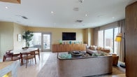 Located in the heart of Downtown Los Angeles, this refined single-floor space at 221 S Grand Ave offers a thoughtfully designed interior defined by warm wood paneling, light stone tile flooring, and an abundance of natural light drawn in through large windows framing expansive city views. The open-concept main living area is anchored by a curved, upholstered sofa ensemble, a wall-mounted flat-screen TV, and a sleek wood credenza, with direct access to an outdoor terrace. A second, more intimate lounge features rich wood-clad walls, a dramatic crystal chandelier, floor-to-ceiling drapes, and sweeping panoramic views of the Los Angeles skyline. The formal dining room centers around a substantial wood table paired with mid-century style chairs, illuminated by a large dome pendant light and accented by curated artwork. The kitchen and entertaining hub showcases a striking marble-topped island with bar seating, illuminated pendant lights, warm wood cabinetry, mirrored backsplash detailing, and a billiards table nearby—ideal for hosting. Throughout the space, a cohesive palette of warm neutrals, walnut tones, and teal accents creates a polished yet inviting atmosphere suited for both relaxation and sophisticated entertaining.