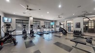 Located at 544 Pitt Greene Connector in Greenville, NC, this single-story fitness center offers a comprehensive layout designed to accommodate a wide range of workout needs. The main exercise floor is expansive and finished with a distinctive speckled tile pattern in alternating light and dark sections, providing clearly defined zones for different activities. Recessed lighting and ceiling fans maintain a bright, comfortable environment throughout.

The cardio area is well-stocked with multiple treadmills, elliptical machines, a stair climber, and a stationary cycle, all positioned along large windows fitted with horizontal blinds that allow natural light to filter in. Wall-mounted televisions are placed throughout for entertainment during workouts. A dedicated strength training section houses a multi-functional cable machine, free weights, and benches, separated from the cardio area by glass partitions that add visual openness while maintaining distinct workout zones.

A separate yoga and stretching studio features herringbone hardwood-style flooring, a striking full-wall peacock feather mural, a mounted flat-screen television, and a bench, creating a calm, focused atmosphere. Vending machines and a hand sanitizer station are conveniently accessible near the entrance, rounding out the practical amenities of this thoughtfully designed facility.