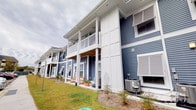 Located at 1500 Coastal Lane in Myrtle Beach, South Carolina, this single-floor residence is part of a well-appointed coastal community designed with classic Lowcountry charm. The exterior immediately captures attention with its bold navy blue lap siding, crisp white board-and-batten accents, and white-railed balconies and covered porches that reflect the relaxed, breezy aesthetic characteristic of the South Carolina coast. A welcoming covered ground-level porch provides an ideal outdoor retreat, complete with rocking chairs that invite residents and guests to enjoy the coastal surroundings. The upper level features a spacious balcony with white railings, extending the outdoor living experience and offering open-air views of the surrounding landscape. The building's design incorporates thoughtful architectural details, including louvered vents, decorative column supports, and a well-maintained lawn that adds a lush, green setting to the property. The community setting suggests shared green spaces and a neighborly atmosphere consistent with planned coastal developments. Overall, the property presents a cohesive blend of traditional coastal architecture and modern residential comfort, making it a distinctive and inviting home in one of South Carolina's most popular beachside destinations.
