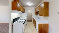 This single-floor, 2-bedroom, 1-bathroom unit offers a straightforward and functional layout designed to maximize every square foot of its compact footprint. The combined kitchen and living area forms the heart of the space. The living room is generously proportioned with soft carpeting underfoot, a textured ceiling, and a wide window framed in warm wood trim that fills the room with natural light. A wall-mounted air conditioning unit and baseboard heaters ensure year-round comfort.

The galley-style kitchen is efficiently arranged with ample warm-toned wood cabinetry lining both walls, providing substantial storage. White appliances including a gas range and refrigerator, a double-basin sink, and light vinyl flooring complete the functional cooking space. A hallway connects the kitchen to the rest of the unit.

The primary bedroom is a spacious, light-filled room featuring carpeted flooring, a wide double window with wood trim, and baseboard heating. The second bedroom is more intimate but equally well-appointed, with carpeting, white walls, and a natural wood-tone door. The bathroom features a clean, classic white tile surround enclosing a full bathtub. A carpeted hallway with a sliding-door closet ties the layout together, adding useful storage near the entryway.