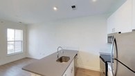 This single-floor space showcases a clean, modern aesthetic throughout its open layout. The kitchen serves as a central highlight, featuring a streamlined kitchen island or peninsula topped with smooth, taupe-toned quartz-style countertops. A stainless steel undermount sink with a contemporary pull-down faucet adds both style and functionality. White shaker-style cabinetry with simple hardware completes the kitchen's crisp, minimalist look, while stainless steel appliances contribute to a cohesive, modern finish.

The flooring throughout is a warm-toned light wood laminate or luxury vinyl plank, lending a sense of warmth that contrasts nicely with the bright white walls and ceiling. Recessed lighting in the ceiling provides even, clean illumination, and a ceiling-mounted ventilation vent speaks to thoughtful utility planning within the space.

The walls are painted a bright, neutral white, amplifying the sense of openness and allowing natural light to reflect freely throughout the room. The overall ambiance is fresh, uncluttered, and move-in ready, making it an ideal canvas for a range of interior styles. This space is well-suited for those seeking a functional yet stylish modern home environment.