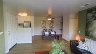 This single-floor space is designed around an open-concept floor plan that seamlessly connects the living, dining, and kitchen areas into one cohesive environment. Hardwood-style flooring runs throughout, lending warmth and visual continuity across the space.

The kitchen is positioned to one side and features white cabinetry, a refrigerator, and a raised breakfast bar or counter that partially separates it from the rest of the living area without disrupting the open feel. A pass-through design keeps the kitchen visually connected to the adjoining spaces.

The dining area sits centrally within the open layout and is anchored by a dark-toned rectangular dining table with matching bench seating. Two botanical-themed wall art pieces frame the dining space, adding a natural, decorative element to the neutral walls.

A ceiling fan with a light fixture is centrally mounted, providing both air circulation and ambient lighting. Built-in shelving on one wall offers functional display space, while the living area accommodates seating and greenery, adding life and color to the room. The overall ambiance is casual, clean, and practical.