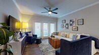 This single-story residence offers a thoughtfully designed open-concept layout that seamlessly connects the main living spaces. The living room serves as a central gathering area, featuring crown molding, wood-style flooring, and a ceiling fan overhead. French doors flood the space with natural light and provide access to an outdoor area, while a curated gallery wall adds a decorative focal point above the seating area.

The kitchen is outfitted with warm-toned cabinetry, black appliances including a refrigerator and range, and a peninsula with a marble-style countertop and integrated sink. Track lighting illuminates the workspace, and the open sightlines to an adjacent dining area make the layout practical for entertaining.

Both bedrooms are carpeted for comfort and equipped with ceiling fans. One bedroom benefits from large windows with sheer drapery, allowing ample natural light, and includes direct access to a walk-in closet. The other bedroom is generously sized with notable wall space and direct hallway access to the bathroom.

The bathroom features a rich wood-toned vanity with a light countertop, a well-lit mirror, and wood-style flooring that continues the cohesive design language found throughout the unit. Overall, the space presents a clean, contemporary aesthetic with a consistent blue and neutral color palette.