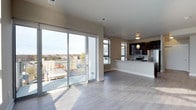 This contemporary multi-floor residence offers a thoughtfully designed open-concept living space bathed in natural light. The main living area features light hardwood-style flooring and a seamless flow toward a large sliding glass door that opens to a private balcony, framing expansive views of the surrounding urban landscape and treetops beyond. The kitchen is anchored by dark cabinetry, pendant lighting, and stainless steel appliances — including a refrigerator, range, and microwave — all centered around a clean-lined counter that doubles as a breakfast bar, blending function with modern style. Neutral toned walls and recessed ceiling vents contribute to the clean, polished aesthetic throughout. One of the most striking features of this residence is the rooftop deck, accessible from the upper floor. This generously sized outdoor space is finished with composite wood decking and enclosed by modern paneled railings, offering breathtaking panoramic views of the city skyline on the horizon and the lush tree canopy below. Whether enjoying morning light or evening city views, this rooftop terrace serves as a remarkable extension of the living space, ideal for both relaxing and entertaining.