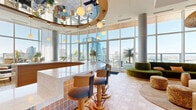 Located at 110 Beech Street in San Diego's vibrant Little Italy neighborhood, this single-floor living space offers a refined and visually striking interior defined by thoughtful architectural design and premium finishes throughout. Soaring double-height ceilings amplify the sense of openness, while an expansive wraparound curtain wall of floor-to-ceiling glass floods the space with abundant natural light and frames panoramic views of the San Diego skyline, waterfront, and outdoor terrace with a pool beyond.

The living area is anchored by a sleek, modern fireplace integrated into a fluted wall panel beneath a wall-mounted screen, complemented by plush velvet seating in earthy olive tones and woven accent ottomans. Adjacent lounge nooks feature deep sectional sofas set against sheer linen curtains, with the cityscape as a backdrop. Structural concrete columns add an industrial counterpoint to the warm wood and marble accents found throughout.

A curved central bar serves as a focal point, clad in geometric patterned tile and topped with a white stone surface, surrounded by wooden barstools with teal upholstery. Orb pendant lighting cascades from a sculpted curved ceiling detail overhead, lending a dramatic, curated ambiance. Lush tropical greenery and sculptural décor pieces complete the sophisticated, resort-inspired aesthetic of this exceptional San Diego space.
