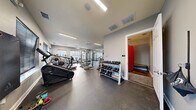 This fitness center offers a comprehensive workout environment designed to accommodate a wide range of exercise needs. The main workout area is generously sized, featuring durable rubber flooring that provides both safety and comfort underfoot. Overhead, linear fluorescent lighting and a ceiling fan ensure the space remains well-lit and ventilated throughout. A full wall of mirrors along one side visually expands the room while allowing users to monitor their form during exercise.

The cardio section is equipped with multiple treadmills and a StairMaster, positioned near large windows fitted with horizontal blinds that allow natural light to filter in. The strength training area includes a multi-station power rack, a cable machine, adjustable benches, and a well-stocked dumbbell rack with a comprehensive range of free weights. The neutral gray walls and clean white trim contribute to a functional yet polished aesthetic throughout the main floor.

An adjoining auxiliary room, accessible through a dedicated doorway, provides additional space and features a striking red heavy bag, a cushioned bench, and a richly toned concrete floor — ideal for martial arts, stretching, or cooldown activities. Together, the two areas form a complete, thoughtfully arranged fitness facility suited for diverse training routines.