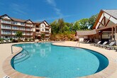 This outdoor amenity space is designed to deliver a resort-like experience, centered around a generously sized freeform swimming pool with crystal-clear blue water and a smooth surrounding deck finished in warm beige concrete, providing ample space for sunbathing. Rows of lounge chairs line the pool's perimeter, offering plentiful seating for residents and guests. A charming covered pavilion anchors one side of the pool area, featuring soaring vaulted ceilings with exposed wood beams, ceiling fans, and large glass doors that open directly to the pool deck. Inside the pavilion, comfortable wicker lounge chairs with plush cushions and a coffee table create an inviting social gathering space, complemented by a ping-pong table for recreation. An attached kitchenette area features white cabinetry with stone countertops, a stainless steel sink, and a stacked stone accent wall, making outdoor entertaining convenient. A second, more intimate pool is also part of the complex, surrounded by lounge seating and shaded umbrella tables, ideal for quieter relaxation. The entire outdoor space is enclosed by wrought iron fencing and framed by mature trees, providing both privacy and a natural, serene ambiance. Tudor-style residential buildings serve as a picturesque backdrop, enhancing the community's cohesive architectural character.