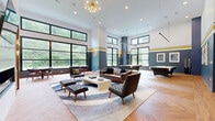 Located at 201 Munson Street in New Haven, CT, this single-floor amenity space offers a generously sized, modern retreat designed for recreation and socializing. The interior is defined by soaring double-height ceilings, floor-to-ceiling black-framed windows, and a bold design palette of slate blue and white walls accented by a distinctive yellow stripe — lending the space a contemporary, energetic character throughout.

The main lounge area features mid-century modern leather seating arranged around a central coffee table, anchored by a statement sputnik chandelier overhead. Herringbone-patterned hardwood flooring flows seamlessly across the space, unifying its various zones. A full billiards table and an air hockey table provide additional entertainment options, while a dedicated golf simulator room — complete with a projection screen, turf mat, and darkened enclosure — offers an immersive experience for enthusiasts.

A wet bar and café-style dining area include subway tile walls, pendant lighting, navy cabinetry, and industrial-style high-top tables and stools, ideal for casual gatherings. A glass-partitioned flex room provides a more private setting for meetings or events. Large windows throughout frame lush green outdoor views, filling the space with abundant natural light.