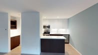 This single-floor residence at 3700 10th Avenue, Unit 1K, showcases a modern design aesthetic with clean lines and a neutral color palette. The kitchen serves as a central highlight of the space, combining functionality with contemporary style. It features floor-to-ceiling white cabinetry that maximizes storage, complemented by dark espresso-toned lower cabinets and a peninsula that provides additional prep space and casual seating potential. Stainless steel appliances, including a gas range, built-in microwave, and dishwasher, are seamlessly integrated into the design. A white tile backsplash adds texture while maintaining the clean, minimalist look. Recessed lighting illuminates the kitchen evenly, enhancing the bright atmosphere created by the light-toned walls. Light hardwood-style flooring flows throughout, adding warmth and continuity to the space. The open layout between the kitchen and living area promotes a sense of spaciousness, making the unit feel airy and well-connected. The overall ambiance is modern, clean, and move-in ready, with thoughtful design choices that balance practicality with contemporary aesthetics.