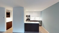 This single-floor residence at 3700 10th Avenue, Unit 1K, showcases a modern design aesthetic with clean lines and a neutral color palette. The kitchen serves as a central highlight of the space, combining functionality with contemporary style. It features floor-to-ceiling white cabinetry that maximizes storage, complemented by dark espresso-toned lower cabinets and a peninsula that provides additional prep space and casual seating potential. Stainless steel appliances, including a gas range, built-in microwave, and dishwasher, are seamlessly integrated into the design. A white tile backsplash adds texture while maintaining the clean, minimalist look. Recessed lighting illuminates the kitchen evenly, enhancing the bright atmosphere created by the light-toned walls. Light hardwood-style flooring flows throughout, adding warmth and continuity to the space. The open layout between the kitchen and living area promotes a sense of spaciousness, making the unit feel airy and well-connected. The overall ambiance is modern, clean, and move-in ready, with thoughtful design choices that balance practicality with contemporary aesthetics.
