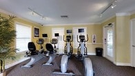 This fitness center is designed to provide a functional and comfortable workout environment. The space features warm yellow walls accented by crisp white crown molding and baseboards, lending a clean and inviting ambiance. The room is carpeted throughout and illuminated by a combination of natural light streaming through multiple windows with white plantation shutters and track lighting mounted on the ceiling.

The cardio equipment selection is well-rounded, including elliptical trainers, upright stationary bikes, and a recumbent bike, each equipped with its own personal monitor screen for entertainment during workouts. The machines are arranged to make efficient use of the floor space while maintaining a comfortable, open feel. A fire extinguisher station is conveniently installed on the wall near the exit, reflecting attention to safety standards.

Decorative touches, including a framed athletic poster on one wall, contribute to the motivating atmosphere of the room. Overhead, a security camera ensures a safe environment for users. The single-floor layout is straightforward and easy to navigate, with a clearly marked exit door. Overall, this fitness center combines practical amenities with a tidy, well-maintained setting suitable for a variety of cardio-focused workout routines.