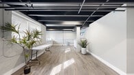 Located at 4645 Pennsylvania Ave in Kansas City's vibrant Westport neighborhood, this shared laundry facility offers a clean, functional, and thoughtfully updated space. The room spans a comfortable footprint and is characterized by its industrial-style open ceiling, finished in black to expose structural beams, ductwork, and piping — a design choice that adds visual character while maintaining a modern aesthetic.

The floor is covered in light wood-look luxury vinyl plank, lending warmth to the space and contrasting effectively with the white-painted concrete block walls and bright white baseboards. Natural light enters through several windows fitted with horizontal blinds, supplementing the overhead lighting and contributing to an open, airy feel.

The laundry equipment is arranged along the perimeter walls, with multiple commercial-grade washing machines and dryers positioned for easy access. Venting infrastructure is neatly routed through the windows, and a folding table provides a practical surface for residents. Decorative potted plants are placed throughout, softening the utilitarian nature of the space. A glass-panel entry door and clearly posted informational signage complete the well-organized, user-friendly environment.