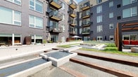 This contemporary outdoor amenity space is defined by its striking courtyard design, seamlessly integrated between the wings of a modern multi-story residential building. The architecture of the surrounding structure features a compelling mix of dark brick cladding, vertical metal panel facades, and private balconies that frame the open-air environment on multiple levels.

At the heart of the courtyard, a series of cascading water features serve as the central focal point, with concrete-edged reflecting pools and gentle water flows creating a calming ambiance. Gravel pathways and broad concrete steps with corten steel accents guide visitors through the space, connecting distinct zones for relaxation and gathering. Raised planter beds filled with native plantings and mulched garden areas introduce natural greenery throughout, softening the clean geometric lines of the hardscape.

Comfortable seating areas with red accent furnishings and wooden benches with concrete surround offer inviting spots to gather or unwind. A separate rooftop terrace extends the outdoor experience further, providing open views of the surrounding urban skyline and mature tree canopy beyond the building perimeter. Abundant natural light floods the entire space throughout the day, making this a vibrant and welcoming outdoor retreat.