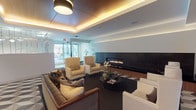 This elegantly designed single-floor lounge space combines modern architectural detailing with a warm, inviting atmosphere. The ceiling is a standout feature, showcasing rich wood paneling framed by a recessed tray with integrated ambient strip lighting that casts a warm golden glow throughout the room. Large pendant light fixtures complement this overhead design, adding both function and visual interest.

Dominating one wall is a wide, linear gas fireplace set within a sleek dark surround with a wood-accented base, creating a striking focal point for the seating area. The fireplace is flanked by a dark, smooth wall panel that reinforces the contemporary aesthetic.

The lounge is furnished with a curated arrangement of plush, neutral-toned sofas and wingback armchairs, accented with decorative throw pillows and a round black coffee table styled with fresh orchids and reading materials. The dark flooring grounds the space and contrasts effectively with the light upholstery.

Natural light filters in through large glazed doors or windows visible at the far end of the space, connecting the interior to an outdoor or corridor area. The overall ambiance is refined and comfortable, suited for relaxed social gatherings or quiet lounging.