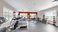 Located at 801 E Brannon Rd in Nicholasville, Kentucky, this fitness center offers a comprehensive workout environment within a single, open-plan space. The room is defined by a bold design aesthetic, featuring a striking red accent wall that contrasts with neutral gray walls and a clean white ceiling fitted with recessed lighting. Durable rubber flooring spans the entire area, providing a safe and resilient surface suited for a wide range of exercise activities.

The cardio zone is generously equipped with multiple treadmills, elliptical trainers, and a recumbent cycle, all positioned along the windowed walls to take advantage of natural light and outdoor views of the surrounding pool area and greenery. A wall-mounted television accompanies the cardio section for entertainment during workouts.

The strength training area features a multi-station cable machine, an adjustable weight bench, and a well-stocked dumbbell rack offering a range of free weights. Medicine balls and stability balls round out the functional fitness offerings. Full-length mirrors are strategically placed to assist with form and to visually expand the space.

Multiple windows fitted with white blinds line the walls, flooding the room with natural light and offering pleasant views of the exterior landscaping. The overall ambiance is energetic yet clean and organized, making this fitness center a practical and inviting facility for residents.