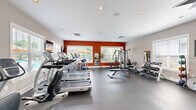 Located at 801 E Brannon Rd in Nicholasville, Kentucky, this fitness center offers a comprehensive workout environment within a single, open-plan space. The room is defined by a bold design aesthetic, featuring a striking red accent wall that contrasts with neutral gray walls and a clean white ceiling fitted with recessed lighting. Durable rubber flooring spans the entire area, providing a safe and resilient surface suited for a wide range of exercise activities.

The cardio zone is generously equipped with multiple treadmills, elliptical trainers, and a recumbent cycle, all positioned along the windowed walls to take advantage of natural light and outdoor views of the surrounding pool area and greenery. A wall-mounted television accompanies the cardio section for entertainment during workouts.

The strength training area features a multi-station cable machine, an adjustable weight bench, and a well-stocked dumbbell rack offering a range of free weights. Medicine balls and stability balls round out the functional fitness offerings. Full-length mirrors are strategically placed to assist with form and to visually expand the space.

Multiple windows fitted with white blinds line the walls, flooding the room with natural light and offering pleasant views of the exterior landscaping. The overall ambiance is energetic yet clean and organized, making this fitness center a practical and inviting facility for residents.