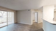 This single-story space is thoughtfully designed with a cohesive, neutral color palette of warm beige and gray tones throughout. Wood-style plank flooring runs continuously across the main living areas and bedrooms, providing a clean, unified aesthetic. Textured ceilings are present throughout the unit.

The living area is open and well-proportioned, featuring a front entry door and a sliding glass door that connects to an outdoor balcony, allowing natural light to filter into the space. A ceiling fan is installed overhead.

The kitchen is positioned adjacent to the living area and features a raised peninsula that creates a natural visual divider while maintaining an open-concept feel. White shaker-style cabinetry, granite-look countertops, and stainless steel appliances — including a refrigerator, microwave, and dishwasher — equip the kitchen efficiently. Light tile flooring distinguishes this area.

The bedroom is generously sized, with a ceiling fan, a window with shuttered blinds providing natural light, and access to a walk-in closet. The connected bathroom includes a white vanity with granite-look countertops, a large mirror, and a full tub-shower combination with tile surround.