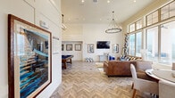 Located at 5771 Oakwood Rd NW in Huntsville, Alabama, this community clubhouse lounge is a thoughtfully designed gathering space that blends modern farmhouse aesthetics with inviting, upscale comfort. The single-floor layout encompasses a generously sized living area anchored by striking herringbone-patterned wood-look flooring that runs throughout the entire space.

Soaring ceilings create an airy, open atmosphere, accentuated by two statement chandelier-style light fixtures — one candelabra-inspired ring pendant and one linear multi-bulb fixture — that add a refined industrial touch. Shiplap-clad accent walls in crisp white serve as a clean backdrop for a wall-mounted flat-screen television, flanked by framed artwork, and a console table with decorative accents.

The main seating area features rich cognac leather sofas arranged around a solid wood coffee table, layered over a bold geometric area rug in mustard, blue, and gray tones. Tall floor-to-ceiling windows dressed with colorful geometric drapery panels flood the space with natural light and provide direct views of the outdoor pool and courtyard beyond.

A dedicated billiards nook offers a full-size pool table with a teal felt surface, complemented by a built-in banquette bench upholstered in navy blue and framed wall panels. Recessed lighting throughout ensures even, well-lit ambiance. The overall design balances playful color with sophisticated architectural detail.