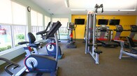 Located at 3857 Flowerfield Rd in Norfolk, Virginia, this fitness center offers a well-equipped, single-floor workout facility designed to support a full range of exercise routines. The space is thoughtfully laid out to accommodate both cardiovascular training and strength work, with clearly defined zones for each.

The cardio area features a row of modern elliptical trainers and treadmills, each equipped with individual display screens for entertainment and workout tracking. A bold yellow accent wall provides an energizing backdrop, while mounted flat-screen televisions line the wall for added convenience. Natural light filters in through roller shade-equipped windows, offering views of the surrounding greenery and creating a welcoming atmosphere.

The strength training section is equally well-appointed, housing a multi-station cable machine, a leg extension/curl bench, and a full dumbbell rack with an accompanying weight bench and wall-length mirror. A water station is conveniently positioned nearby. Additional storage cubbies and accessory holders are integrated throughout the space for organizational convenience.

The flooring throughout is durable, textured carpet suited for gym use, and ceiling fans assist with air circulation. The neutral gray walls, bright overhead lighting, and clean layout contribute to a functional and comfortable environment for all fitness levels.