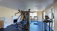 This single-floor fitness center is designed to support a comprehensive workout experience within a comfortable, well-maintained environment. The space features a multi-station cable and weight machine — a Hoist H2200 — positioned prominently at the center, offering a wide range of strength training exercises. Complementing this is a set of treadmills arranged along the far wall, providing dedicated space for cardiovascular training. Natural light filters in through the windows, creating a bright and energizing atmosphere. The flooring consists of dark rubberized material, practical and appropriate for a fitness setting. A notable architectural detail is the classic white-painted fireplace with a brick hearth along one wall, adding a distinctive and welcoming character to what might otherwise be a purely utilitarian space. The ceiling features a textured finish with recessed and surface-mounted lighting, ensuring the room is well-lit throughout. The walls are painted in a soft, neutral tone, contributing to the overall clean and focused ambiance. This fitness center balances functionality with comfort, making it a practical and inviting space for daily exercise routines.
