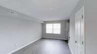 This two-story space presents a clean, contemporary aesthetic defined by a consistent and cohesive design palette. Light gray walls pair seamlessly with smooth white ceilings and light-toned hardwood-style flooring, creating a bright and neutral canvas throughout. The main living area is open and airy, benefiting from natural light streaming through a large window that illuminates the space and highlights the clean lines of the interior. White baseboards and trim provide a crisp contrast to the soft gray walls, adding a polished finish to each room. Ceiling-mounted light fixtures and recessed HVAC vents are thoughtfully integrated, maintaining the clean, uncluttered look of the space. A glimpse into the secondary areas reveals a continuation of the same refined design language, with dark cabinetry visible in what appears to be a kitchen area, offering a striking contrast to the otherwise light interior. Interior doors are finished in white, contributing to the overall sense of brightness and openness. The layout flows naturally across two floors, offering functional separation of spaces while maintaining a unified design throughout. This well-maintained space is move-in ready and offers a versatile, modern interior suited to a variety of uses.