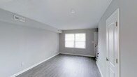 This two-story space presents a clean, contemporary aesthetic defined by a consistent and cohesive design palette. Light gray walls pair seamlessly with smooth white ceilings and light-toned hardwood-style flooring, creating a bright and neutral canvas throughout. The main living area is open and airy, benefiting from natural light streaming through a large window that illuminates the space and highlights the clean lines of the interior. White baseboards and trim provide a crisp contrast to the soft gray walls, adding a polished finish to each room. Ceiling-mounted light fixtures and recessed HVAC vents are thoughtfully integrated, maintaining the clean, uncluttered look of the space. A glimpse into the secondary areas reveals a continuation of the same refined design language, with dark cabinetry visible in what appears to be a kitchen area, offering a striking contrast to the otherwise light interior. Interior doors are finished in white, contributing to the overall sense of brightness and openness. The layout flows naturally across two floors, offering functional separation of spaces while maintaining a unified design throughout. This well-maintained space is move-in ready and offers a versatile, modern interior suited to a variety of uses.