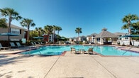 This outdoor pool area in Jacksonville, Florida offers a well-appointed resort-style retreat designed for relaxation and recreation. At the heart of the space is a generously sized swimming pool with crystal-clear blue water, surrounded by a broad concrete sundeck that accommodates multiple seating arrangements, including lounge chairs and umbrella-shaded dining tables ideal for outdoor gatherings.

A standout wooden pergola provides a covered lounge area furnished with comfortable seating and a brick fire pit, creating a cozy atmosphere for evening socializing. Tall, mature palm trees are scattered throughout the property, lending a distinctly tropical character and natural shade across the deck. Lush manicured hedges and colorful tropical plantings frame the perimeter, enhancing privacy and visual appeal.

A charming gazebo-style structure anchors one end of the pool area, while broad brick-accented steps and raised planters connect the pool deck to the adjacent clubhouse building. The clubhouse features traditional architecture with arched windows, a covered entryway, and a wood pergola overhead, adding architectural elegance to the outdoor environment. The entire space benefits from abundant Florida sunshine, open skies, and views of the surrounding residential community, creating a vibrant and inviting outdoor amenity area.