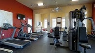 Located at 5300 Patron Place in Summerville, South Carolina, this fitness center is a well-appointed, single-floor exercise facility designed to support a full range of workout routines. The space features a bold accent wall in deep red, contrasting with warm yellow walls that create an energizing yet welcoming atmosphere. Durable rubber flooring covers the workout area, providing both safety and comfort during exercise.

The cardio zone is equipped with multiple treadmills and an elliptical machine, all positioned to take advantage of the natural light streaming in through several windows fitted with classic blinds. A wall-mounted flat-screen television is conveniently placed for entertainment during cardio sessions. A ceiling fan overhead ensures proper air circulation throughout the space.

Strength training is well-supported with a dedicated cable machine and a weight bench, providing users with a versatile range of resistance exercises. Beyond the fitness floor, a wrought iron railing separates an adjacent area visible in the background, suggesting a connected lounge or social space with additional seating and decor. The overall design balances function and aesthetics, making this fitness center a practical and inviting amenity for residents of the community.