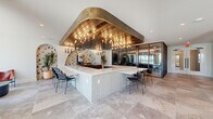 This expansive single-floor space blends contemporary and eclectic design across a generously sized layout. Large-format stone tile flooring flows continuously throughout, unifying the distinct functional zones. The central focal point is a substantial bar counter clad in light vertical tile, topped with a white stone surface and surrounded by seating. Above it, a dramatic recessed ceiling feature combines warm wood planking with a bold bronze surround and striking candelabra-style chandeliers, creating an eye-catching architectural statement.

The lounge area is anchored by a full dark brick fireplace wall with an integrated television, complemented by a curated mix of seating and a woven area rug. Floor-to-ceiling glass walls and sliding doors allow abundant natural light and direct views of an outdoor courtyard. A glass-enclosed conference room with black steel-frame partitions provides a private, transparent workspace visible from the main floor.

The hallway connects zones seamlessly, featuring arched wall niches adorned with bold tropical wallpaper and textured accent walls. Throughout, the design balances industrial elements, organic materials, and artful décor, resulting in a sophisticated and inviting communal environment.