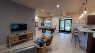 The Resident Activity Center is a single-floor community facility thoughtfully designed to encourage socializing and recreation. Guests are welcomed by an elegant foyer featuring large-format light tile flooring, crown molding, recessed lighting, and a stylish round accent table beneath a statement pendant fixture. The space flows into a spacious open lounge anchored by a cozy fireplace, comfortable seating, and abundant natural light from oversized windows and glass doors that frame views of lush outdoor greenery. A dedicated game room offers a full-size pool table, shuffleboard table, wall-mounted TV, and a built-in display shelving unit, all set against a bold gray accent wall. A bar area features granite countertops with mosaic tile detailing, rich dark cabinetry, globe pendant lighting, and bar stool seating — ideal for casual gatherings. The fully equipped kitchen includes stainless steel appliances, granite countertops, a decorative tile backsplash, and ample dark wood cabinetry. A cozy nook with high-top tables, bar stools, and a wall-mounted TV provides an intimate spot to relax. Throughout the center, consistent tile flooring, neutral tones, curated artwork, and recessed lighting create a polished and inviting ambiance.