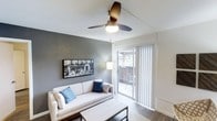 This single-floor residence offers a thoughtfully designed living space with a clean, modern aesthetic. The living room serves as the heart of the home, featuring warm hardwood-style flooring, smooth white ceilings, and a stylish ceiling fan that adds both function and a contemporary touch. An accent wall in a deep charcoal gray creates a bold focal point, complemented by tasteful wall art that enhances the modern atmosphere. Natural light flows generously into the space through large sliding glass doors fitted with vertical blinds, which open directly onto a private outdoor balcony — perfect for enjoying fresh air and outdoor views. The seamless transition between the interior and the balcony gives the living area an open, airy feel. The layout is well-proportioned, comfortably accommodating a sofa and coffee table while maintaining a sense of spaciousness. Overall, the space strikes a balance between comfort and contemporary style, with neutral tones, clean lines, and thoughtful design details that create a welcoming and functional environment.